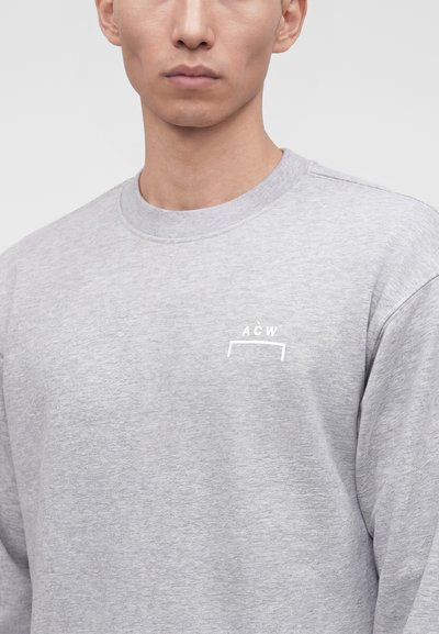 Κοντινό πλάνο ενός ατόμου που φοράει ένα ανοιχτό γκρι φούτερ με στρογγυλή λαιμόκοψη και μικρό λευκό λογότυπο "A C W" στο αριστερό στήθος.