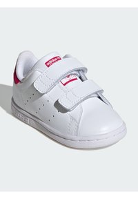White sneakers with two Velcro straps, synthetic leather material, rounded toe, perforated side details, and pink accent on the heel.