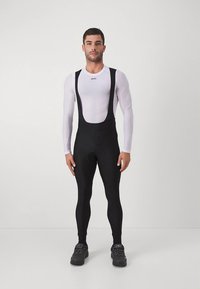 Zwarte fietsbroek met bretels, verstelbare bandjes, vochtafvoerend en een aansluitend ontwerp. Gecombineerd met een wit ondershirt met lange mouwen.