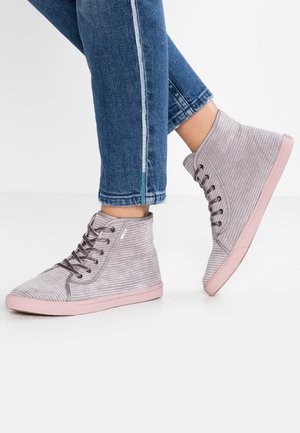 Osoba nosząca jasnoróżowe sznurowane trampki typu high-top w parze z niebieskimi dżinsami, stojąca na białej powierzchni.