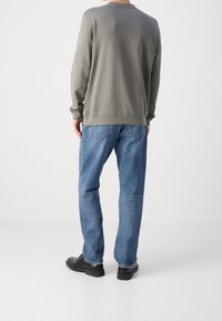 Man som bär en grå sweatshirt med långa ärmar, blå jeans och svarta skor, stående vänd bort från kameran mot en neutral bakgrund.
