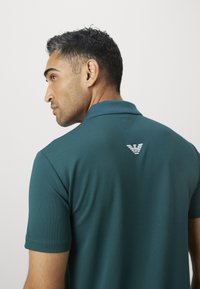 Vyras su trumpais tamsiais plaukais, vilkintis mėlynos-žalios polo marškinėlius su mažu baltu logotipu ant viršutinės nugaros dalies, žiūri į kairę priešais paprastą foną.
