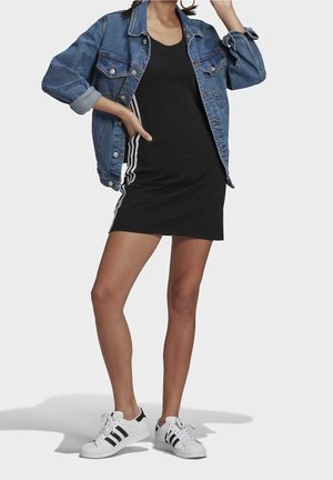 Jeansjacke über einem schwarzen Kleid mit weißen Seitenstreifen, weiße Sneakers mit schwarzen Akzenten, die einen lässigen Schnitt und umgeschlagene Ärmel aufweisen.