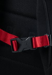 Tela de mochila negra con una correa roja y un hebilla de plástico negro que presenta un logotipo. Superficie texturizada con detalles de costura visibles.