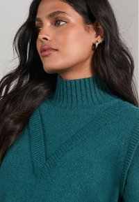 Pulover tricotat de culoare teal, cu guler înalt cu tehnica rib și un design în formă de V. Textură moale și detalii de cusături proeminente pe umeri.