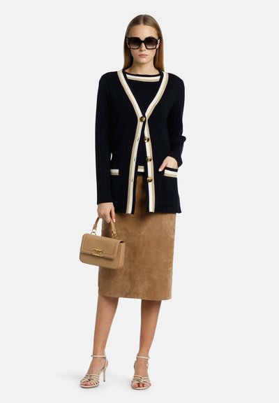 Mörkblå kofta med krämfärgade detaljer och guldknappar, kombinerad med en beige mockaskjorta, sandaletter med remmar och en beige handväska. Modell som bär stora solglasögon.