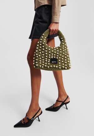 Bolso de ganchillo verde con cuentas blancas en la superficie, con un asa grande y una pequeña etiqueta negra. Combinado con zapatos negros de tacón.