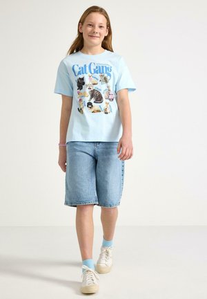 Tyttö kävelee, pukeutuneena vaaleansiniseen Cat Gang -grafiikkapaitaan, polvipitkiin farkkushortseihin, vaaleansinisiin sukkiin ja valkoisiin lenkkareihin.