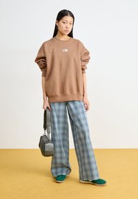 Brązowy, oversize'owy sweatshirt z białym logo, sparowany z szerokimi spodniami w kratę w odcieniach niebieskiego i zielonego. Zielone buty typu slip-on i szara torba.
