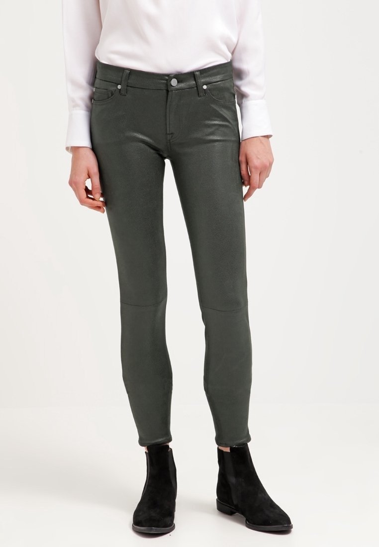Personne portant un pantalon en cuir moulant vert foncé, une chemise blanche à manches longues et des bottines noires, debout devant un fond blanc.