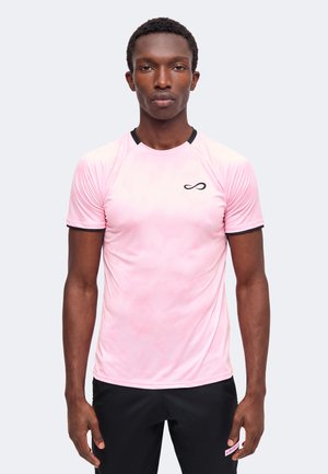 Young man wearing a light pink short-sleeve athletic shirt with black trim and black shorts, standing against a plain light background.