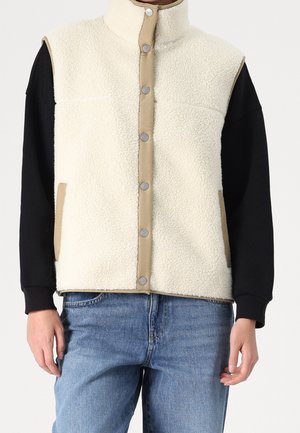 Sherpa fleece vest i cremefarve med tan kant og trykknapper, båret over en sort langærmet skjorte og blå jeans.