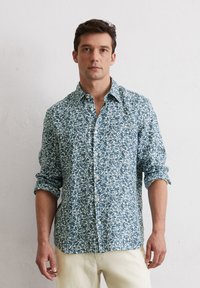 Chemise boutonnée de couleur bleu clair avec un motif de feuilles blanches. Manches longues retroussées, tissu doux, poche plaquée unique sur le côté gauche de la poitrine.