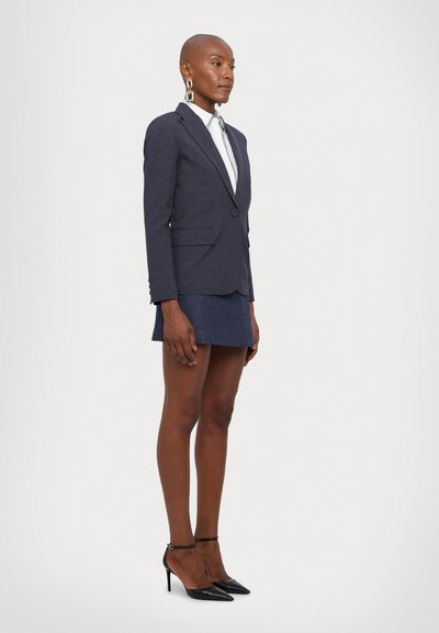 Blazer grigio scuro con due tasche frontali, abbinato a una gonna blu corta. Tacchi a punta neri e grandi orecchini a cerchio completano il look.