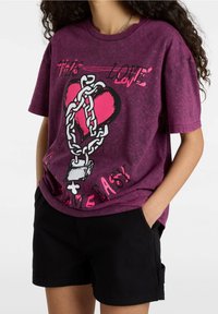 Camiseta de tie-dye morado con un gráfico de un corazón rodeado de cadenas, con texto en rosa y negro. Mangas cortas y corte relajado.