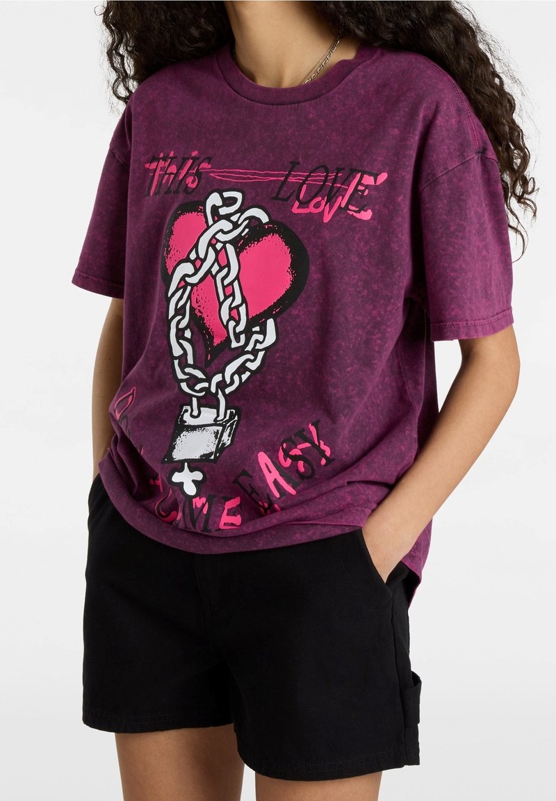 Camiseta de tie-dye morado con un gráfico de un corazón rodeado de cadenas, con texto en rosa y negro. Mangas cortas y corte relajado.