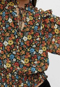 Blommig blus med svart bakgrund, prydd med en mängd färgglada blommor i röd, gul och blå, med samlad midja och volangkrage.
