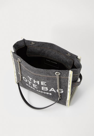 Sac fourre-tout en tissu gris avec le texte blanc "THE TOTE BAG" et "MARC JACOBS". Présente des poignées en chaîne et en cuir, des coutures apparentes et une texture douce.