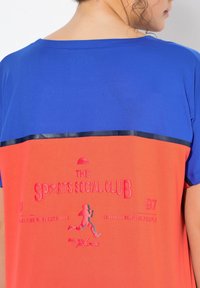 Koszulka sportowa z krótkim rękawem, z niebieską górą i pomarańczowym dołem. Zawiera wytłaczany napis oraz grafikę biegnącej postaci. Napis brzmi "THE SPORTS SOCIAL CLUB."