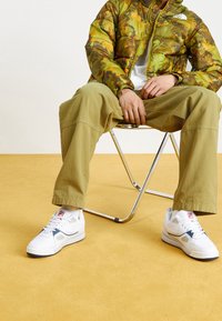 Zeleno-hnědá kamuflážní bunda s polstrováním, bílé a námořnicky modré tenisky s logem, khaki široké kalhoty, sedící na chromové židli.