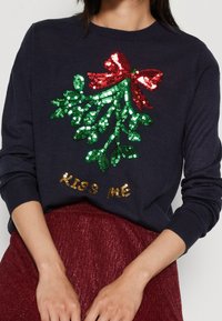 Marineblauwe gebreide trui met een bedel van een misthoornontwerp met een rode strik. Gouden letters spellen "KISS ME" onder de decoratie.
