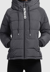 Doudoune grise avec un col montant, fermeture éclair et cordons de serrage. Présente un motif matelassé, des poignets noirs et des poches latérales.