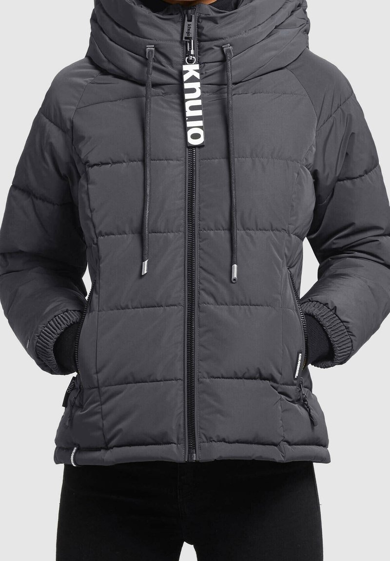 Doudoune grise avec un col montant, fermeture éclair et cordons de serrage. Présente un motif matelassé, des poignets noirs et des poches latérales.