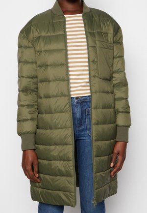 Personne portant une longue veste matelassée vert olive sur une chemise rayée beige et blanche avec un jean bleu, debout devant un fond uni.