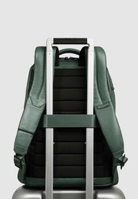 Mochila de cuero verde con correas negras acolchadas, sujetas a un asa plateada de equipaje. Presenta costuras distintivas y una textura en forma de cuadrícula.