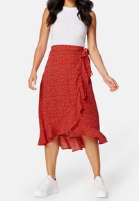 Falda envolvente roja con flores y bajo con volantes, atada en la cintura, combinada con una camiseta ajustada blanca y sneakers blancos.