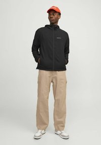 Jachetă neagră cu glugă, fermoar în față, pantaloni cargo bej și adidași albi. Model îmbrăcat cu o pălărie roșie aprinsă cu logo.