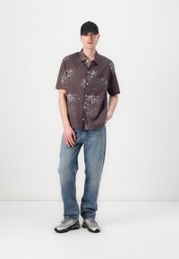 Chemise à manches courtes, boutonnée, de couleur violet foncé avec un motif floral, accompagnée de jeans bleu clair et de chaussures de sport argentées. Casquette noire portée.