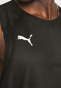 Gros plan d’un débardeur de sport noir en mesh avec un logo Puma blanc sur la poitrine, porté par une personne à la peau foncée.