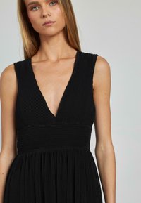 Schwarzes plissiertes Kleid mit tiefem V-Ausschnitt, ärmellosem Design und strukturiertem Stoff. Das Oberteil ist figurbetont und der Rock fließt.