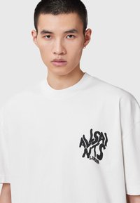 Camiseta de algodón blanca con corte holgado, cuello redondo y estampado gráfico negro "ALLSAINTS LONDON" en el lado izquierdo del pecho. Textura suave.