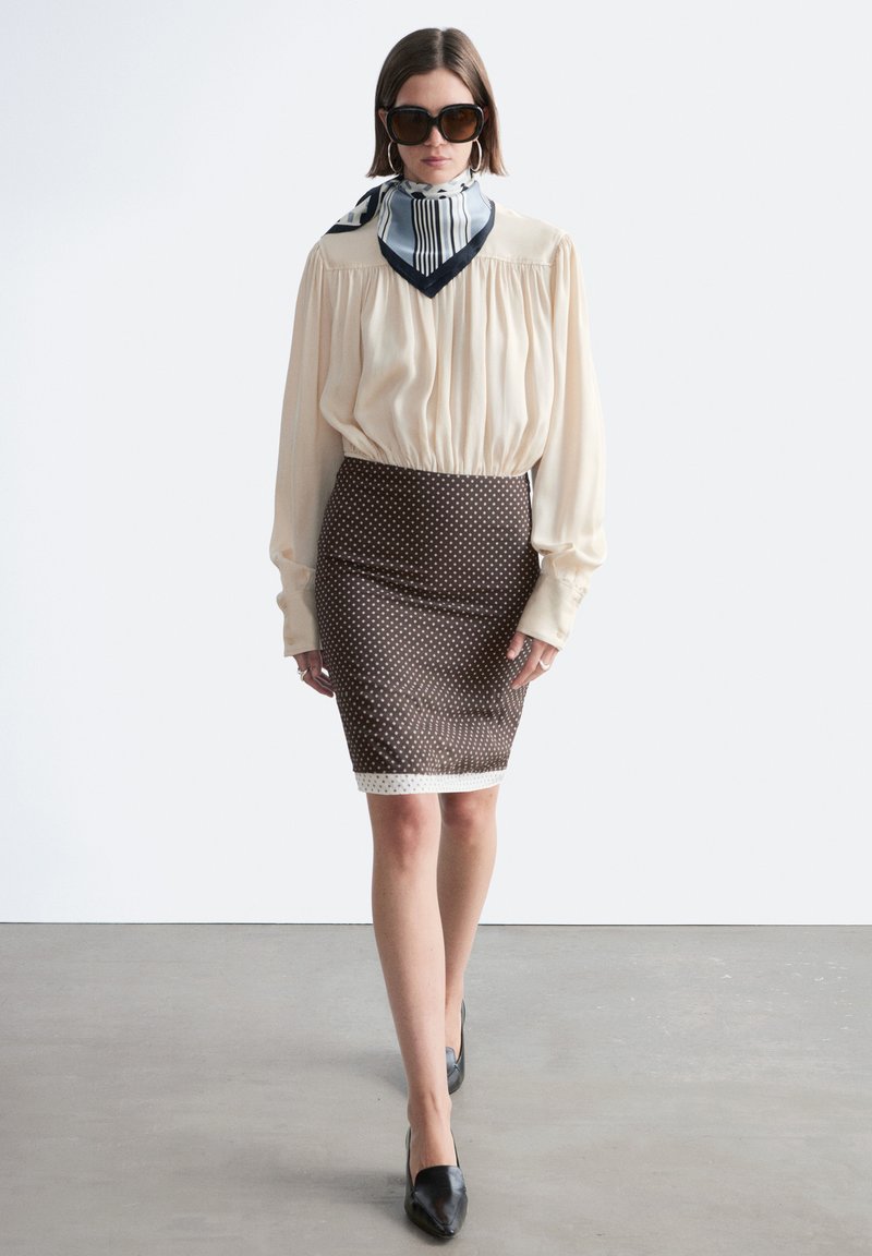 Donna con occhiali da sole scuri oversize, camicetta color crema, gonna a tubino nera a pois, tacchi neri e sciarpa a righe al collo, che cammina in avanti.
