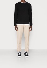 Pull en maille noire avec un col rond blanc, pantalon beige fuselé, et baskets noires à semelles blanches. Tenue décontractée avec des détails contrastants.