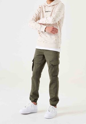 Osoba w beżowej, nakrapianej bluzie z kapturem, oliwkowo-zielonych spodniach typu cargo jogger oraz białych trampkach stoi z założonymi skrzyżowanymi rękami.