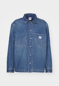 Camicia di jeans di blu medio con chiusura a bottoni, colletto classico, due tasche sul petto e una toppa con etichetta sulla tasca sinistra.
