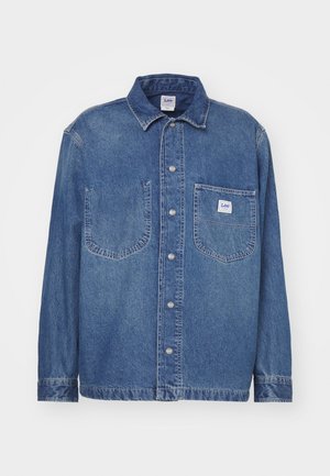 Chemise en denim bleu moyen avec un devant à boutons, un col classique, deux poches poitrine et une étiquette sur la poche gauche.