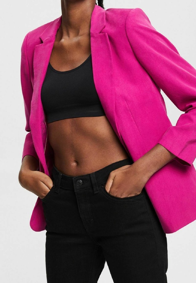 Personne portant un blazer rose vif, un crop top noir et un pantalon noir, les mains dans les poches, sur un fond clair uni.