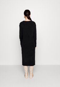 Černé šaty s dlouhým rukávem a volným střihem, pletená tkanina, dosahující ke středu lýtek. V kombinaci s béžovými kozačkami po kolena. Minimální detaily.