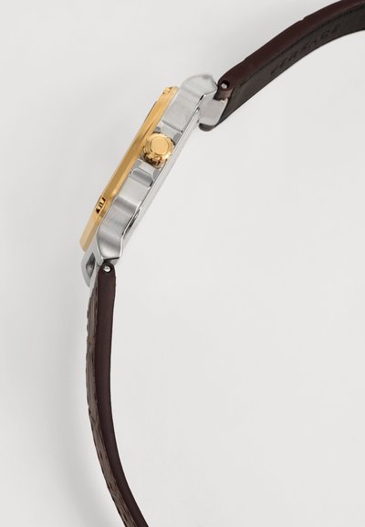 Vue latérale d'une montre-bracelet avec une couronne dorée, un boîtier argenté et un bracelet en cuir brun foncé sur fond blanc uni.