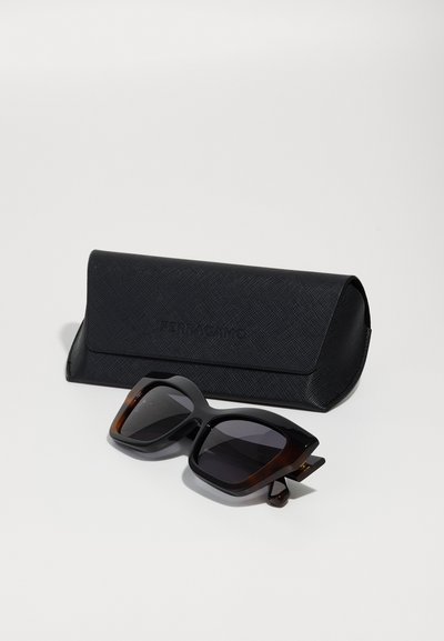 Occhiali da sole in acetato neri con un design squadrato e massiccio e lenti scure. Accompagnati da una custodia nera texturizzata con logo.