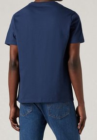 Homme portant un t-shirt uni bleu marine à manches courtes et un jean bleu, vu de dos sur un fond neutre.
