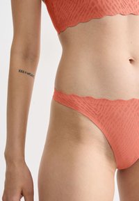 Koraalroze lingerieset met een gegolfde rand. Zachte textuur en geometrisch patroon. Focus op de tailleband en het onderste lichaamsgedeelte, huid zichtbaar.