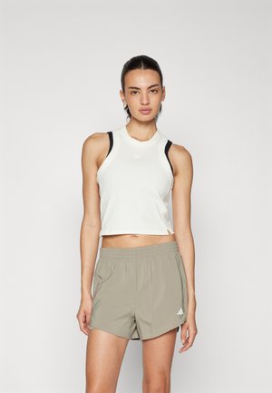 Weißes, geripptes Cropped-Top mit schwarzen Trägern und Logo; kombiniert mit hellolivgrünen Sportshorts mit elastischem Bund und Logo.