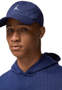 Marineblå cap med hvit Jumpman-logo, båret med en matchende marineblå hettegenser. Mykt stoff, rund skjerm, jevn tekstur, minimale design detaljer.