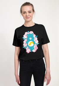 Zwarte t-shirt met een blauwe beerafbeelding die een gele maan vasthoudt, omringd door roze rozen en groene bladeren. Korte mouwen, ronde halsontwerp.