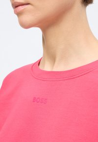 Cou et bas du visage d'une personne portant un t-shirt rose vif avec un logo "BOSS" en relief au centre de la poitrine sur fond blanc.
